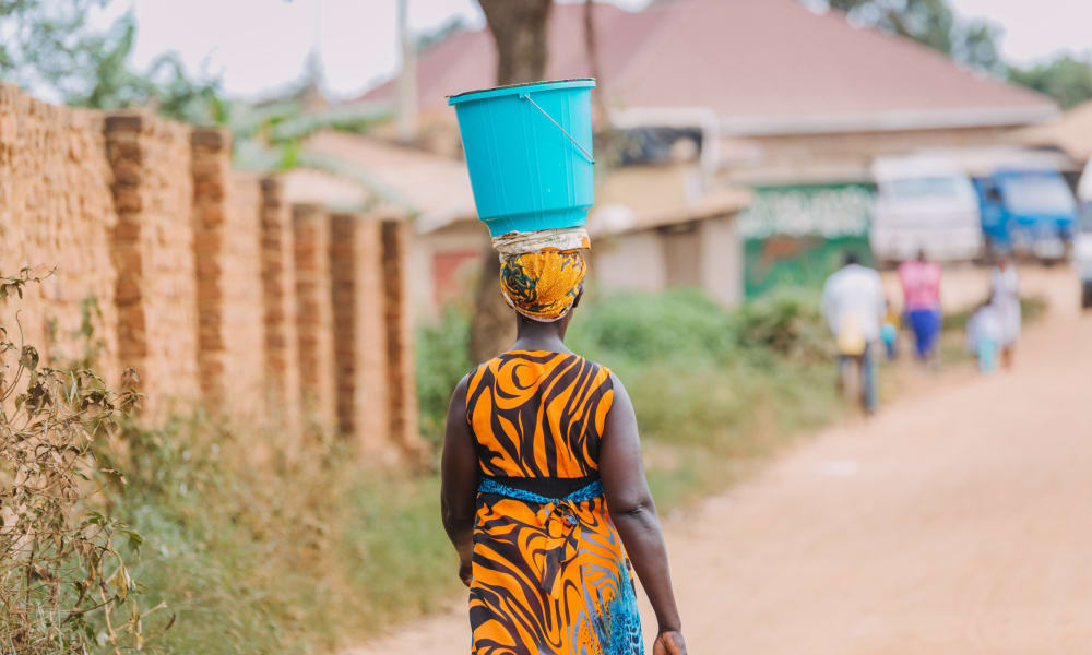 A dark-skinned woman in traditional dress carrying a bucket on her head