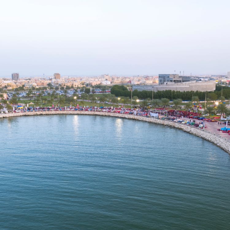 Eine entfernte Luftaufnahme vom Wasser aus von den Oldtimern, die bei der Oldtimershow am Hafen von Dammam entlang der Promenade aufgereiht sind, mit der Stadt Dammam im Hintergrund