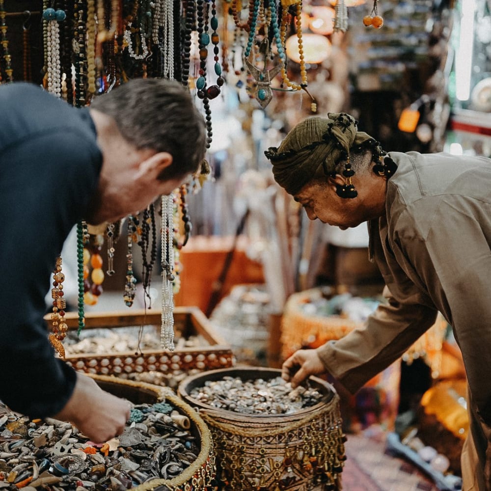 Two people shopping for jewelry and accessories at a busy outdoor market stall with hanging necklaces and bracelets.