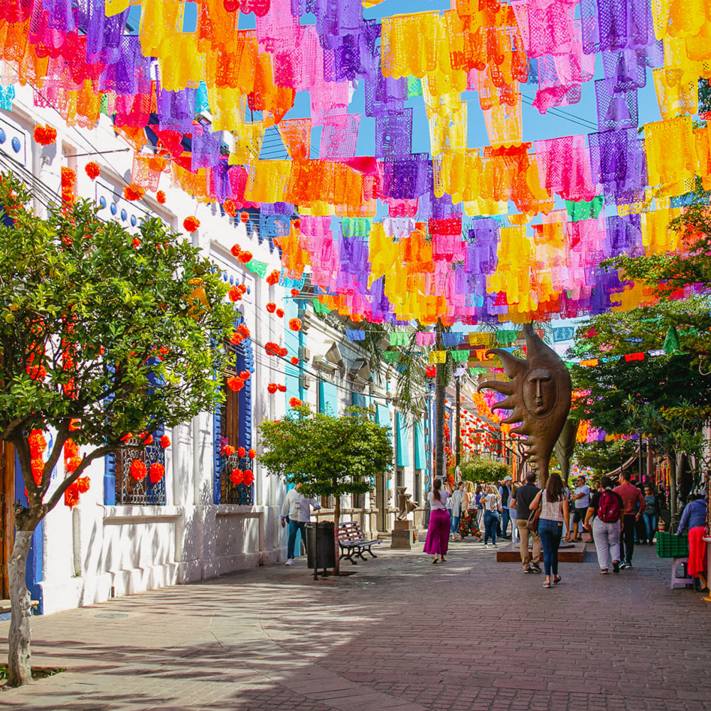 Eine lebendige Straße in Tlaquepaque, Jalisco, Mexiko, geschmückt mit bunten Papierdekorationen, die über Kopf hängen, und Menschen, die entlang der gepflasterten Straße gehen, mit Bäumen und Gebäuden auf beiden Seiten.