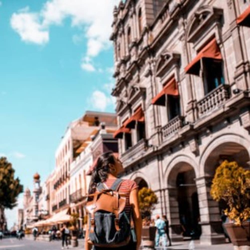 Eine Frau mit Rucksack geht eine Straße in Puebla, Mexiko entlang, gesäumt von historischen Gebäuden mit Bogenfenstern und orangen Markisen unter einem teils bewölkten Himmel.