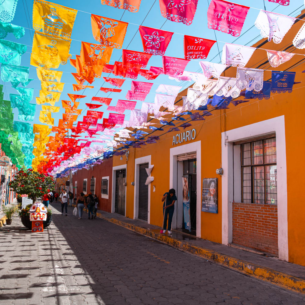 Eine bunte Straße in Atlixco, Puebla, geschmückt mit Reihen farbenfroher Papierfahnen, die über Kopf hängen, und Menschen, die entlang des Kopfsteinpflasterwegs an hell gestrichenen Gebäuden gehen.