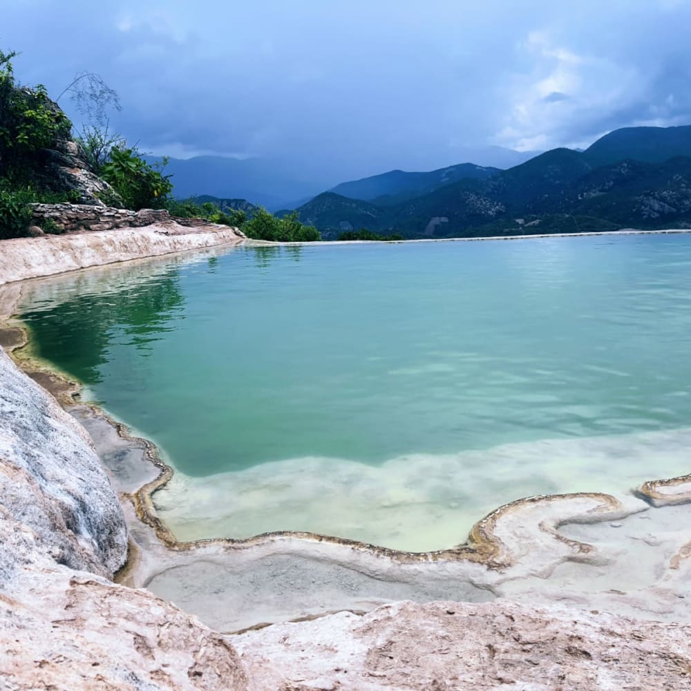 Mineralreiches Naturbecken mit türkisfarbenem Wasser und einzigartigen Sedimentformationen vor einer Kulisse entfernter Bergketten unter einem bewölkten Himmel.