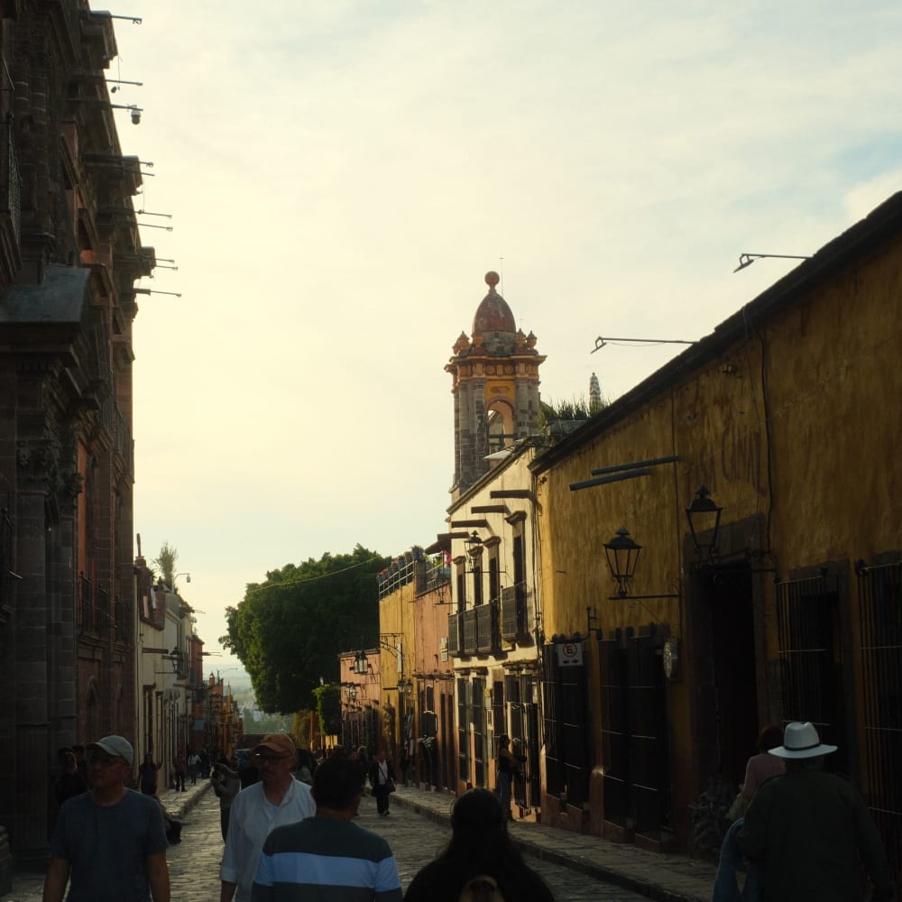 Eine Straßenszene in San Miguel de Allende mit Menschen, die entlang gepflasterter Straßen spazieren, kolonialstillichen Gebäuden und einem Kirchturm im Hintergrund bei frühem Abendlicht.
