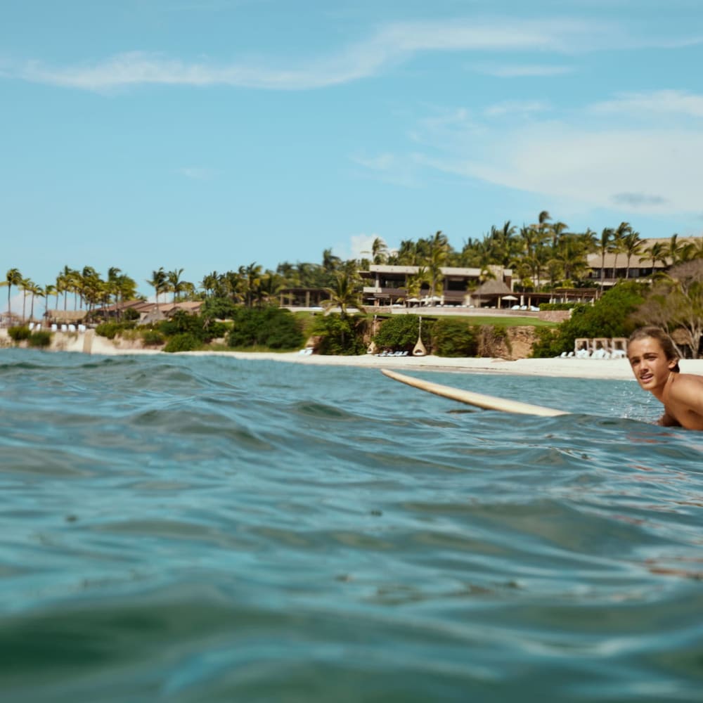 Eine Person liegt auf einem Surfbrett und paddelt im Meer in der Nähe eines tropischen Strandresorts mit Palmen unter einem leicht bewölkten Himmel.