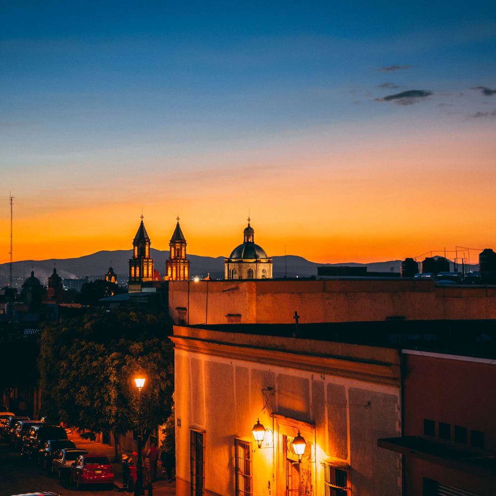 Abendliche Ansicht von Santiago de Querétaro mit beleuchteten Kirchtürmen und Straßenlaternen vor einem farbenfrohen Sonnenunterganghimmel.