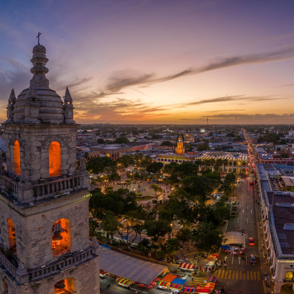 Luftaufnahme von Mérida, Mexiko, mit beleuchteten historischen Kirchtürmen im Vordergrund und einem Stadtplatz mit Bäumen und Gebäuden bei Sonnenuntergang.