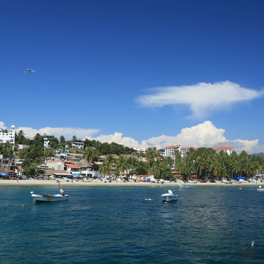 Eine sonnige Küstenansicht von Puerto Escondido, Oaxaca, mit Booten auf blauem Wasser vor einem Sandstrand, der von Palmen und Gebäuden gesäumt ist, unter einem teilweise bewölkten Himmel.