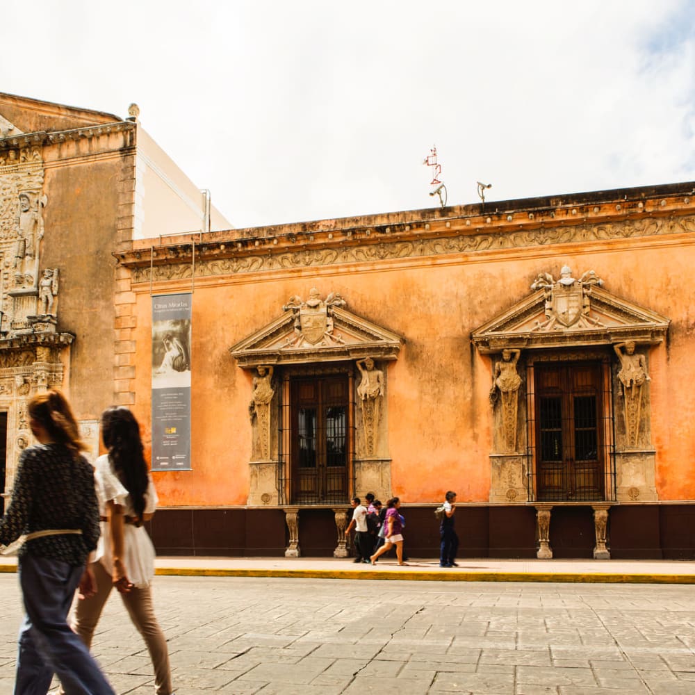 Menschen gehen an einem historischen, reich verzierten Gebäude mit großen Fenstern und einem kunstvoll gestalteten Steineingang in Mérida, Yucatán, Mexiko, vorbei.