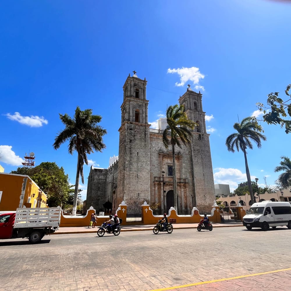 Historische Steinkirche mit zwei hohen Glockentürmen und Palmen davor, an einer sonnigen Straße mit vorbeifahrenden Motorrädern und Fahrzeugen in Valladolid.