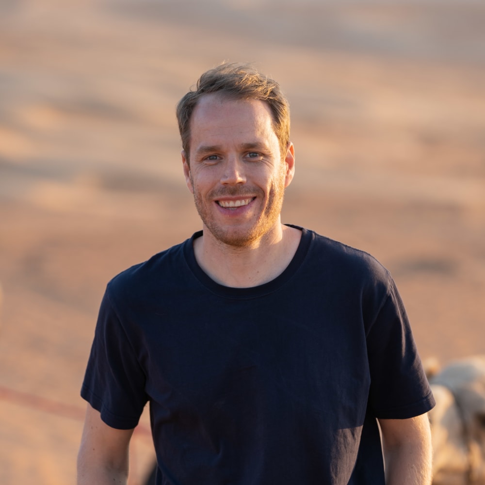 Ein lachender junger Mann mit kurzen Haaren und Bart, trägt ein schwarzes T-Shirt, steht im Freien vor einem sandigen Hintergrund.