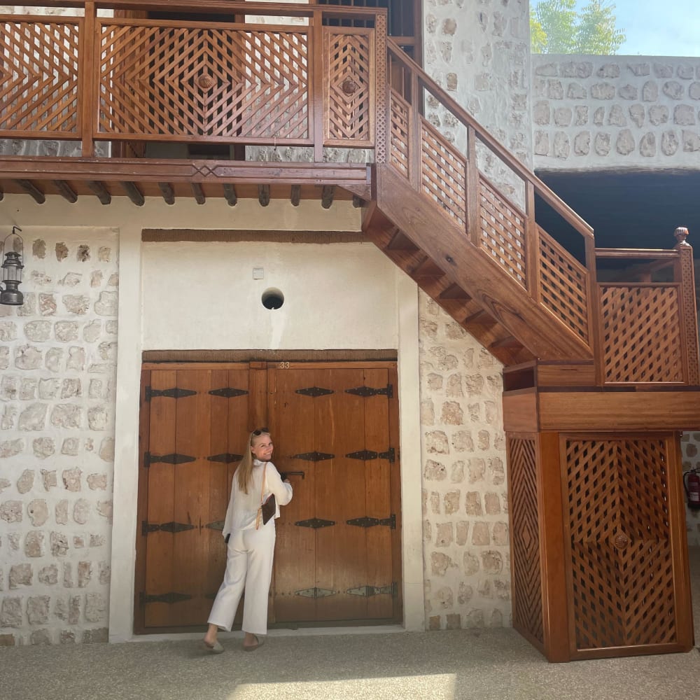 A woman in white pants and a white top stands near large wooden doors in a stone building with a balcony and staircase.