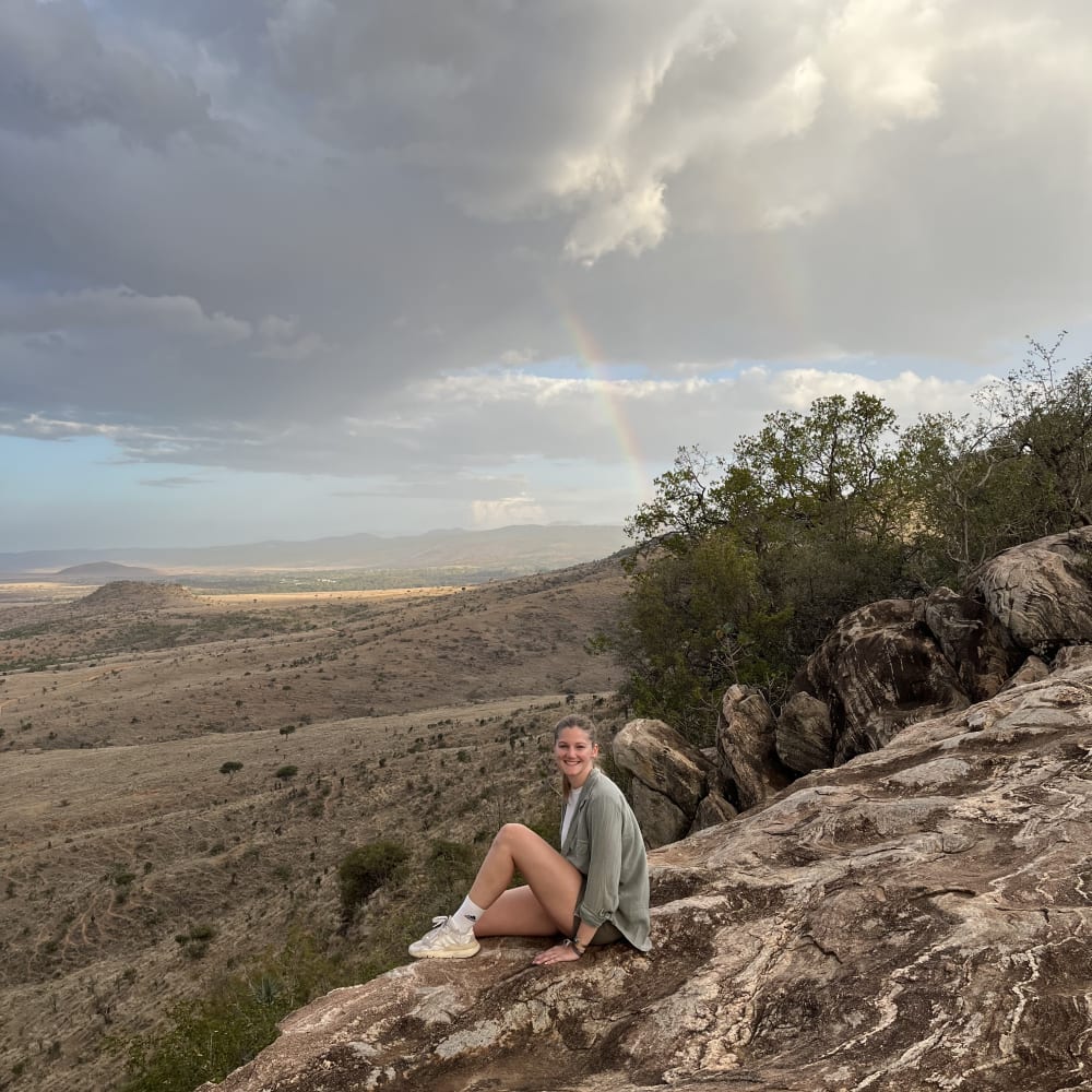 Eine Frau sitzt auf einem großen Felsen, der einen weiten, halbtrockenen Landschaftsausblick bietet, mit Wolken und einem schwachen Regenbogen im Hintergrund.