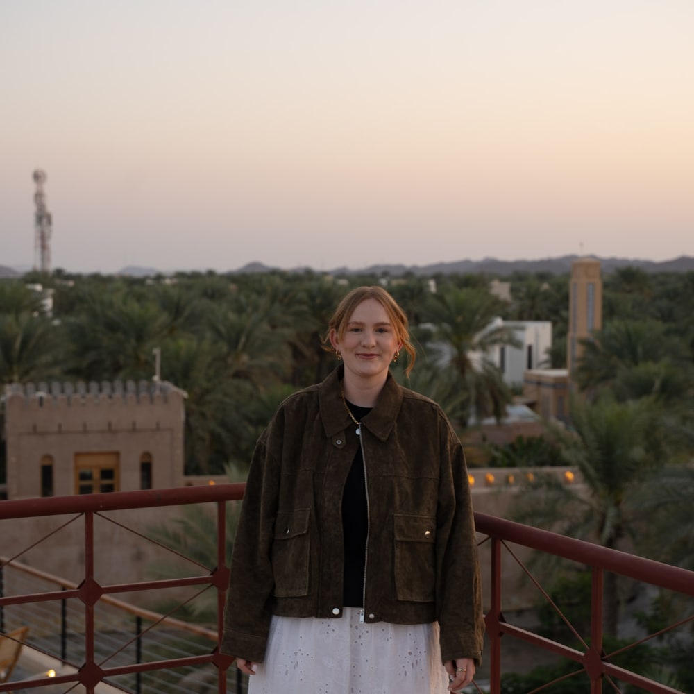 Eine Frau mit hellbraunen Haaren trägt eine dunkle Jacke und einen weißen Rock und steht auf einem Balkon mit Blick auf Palmen und traditionelle Gebäude bei Dämmerung.