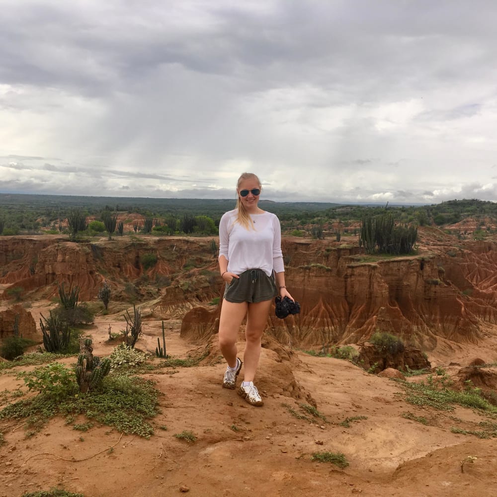 Eine Frau mit Sonnenbrille, weißem Hemd und Shorts steht auf einem sandigen Plateau mit Kakteen und erodierten roten Felsformationen in der Tatacoa-Wüste unter einem bewölkten Himmel.