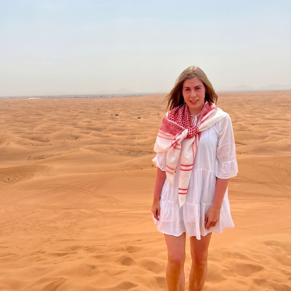 Eine Frau in weißem Kleid und einem rot-weißen Schal steht auf weitläufigen Sanddünen in einer Wüste unter klarem Himmel.