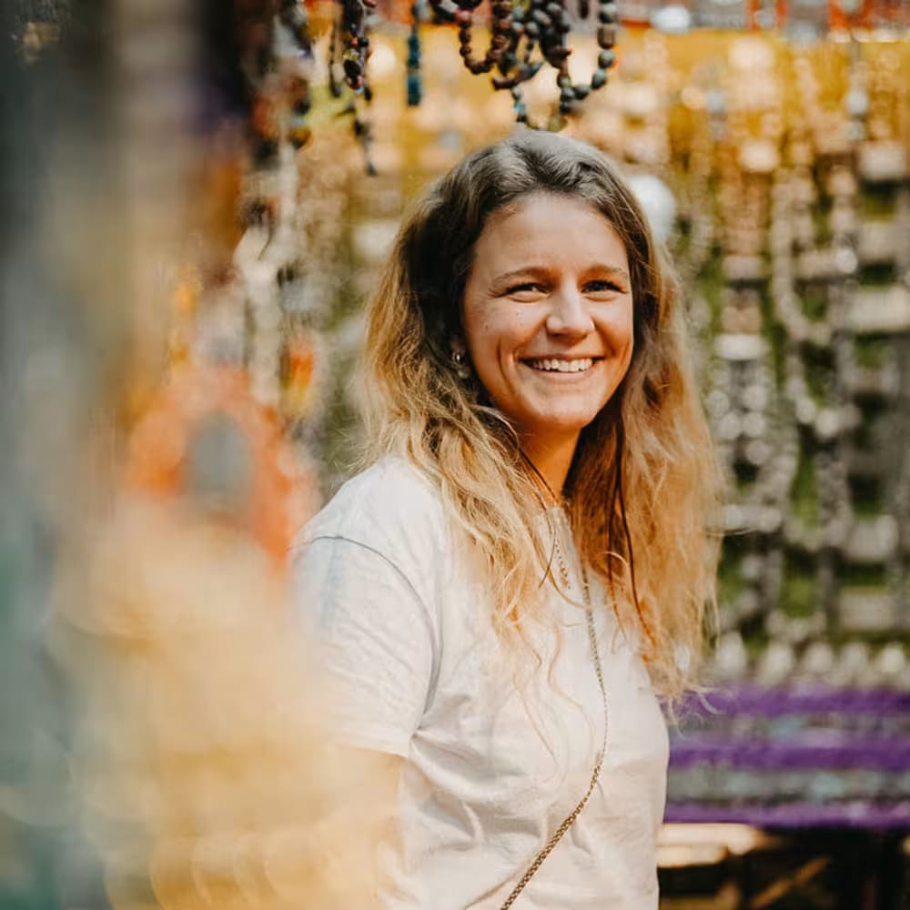A woman with wavy light brown hair is smiling in an outdoor market setting with colorful beads and decorations hanging above.