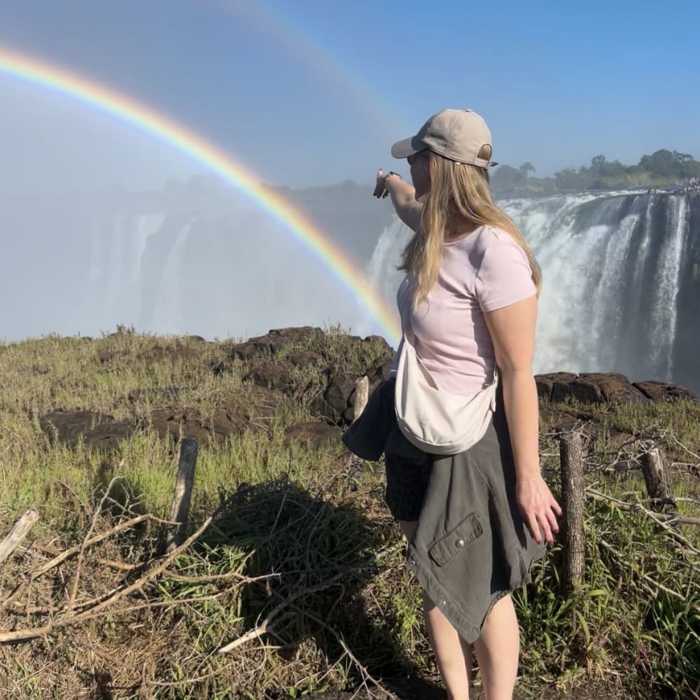 A woman wearing a cap and light pink shirt points towards a vibrant double rainbow near Victoria Falls, with cascading water and mist in the background.