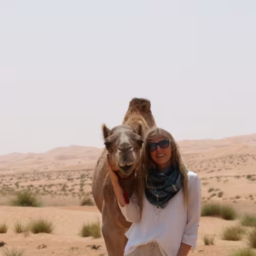 A woman wearing sunglasses stands smiling next to a camel in a desert landscape with sandy dunes and sparse vegetation.