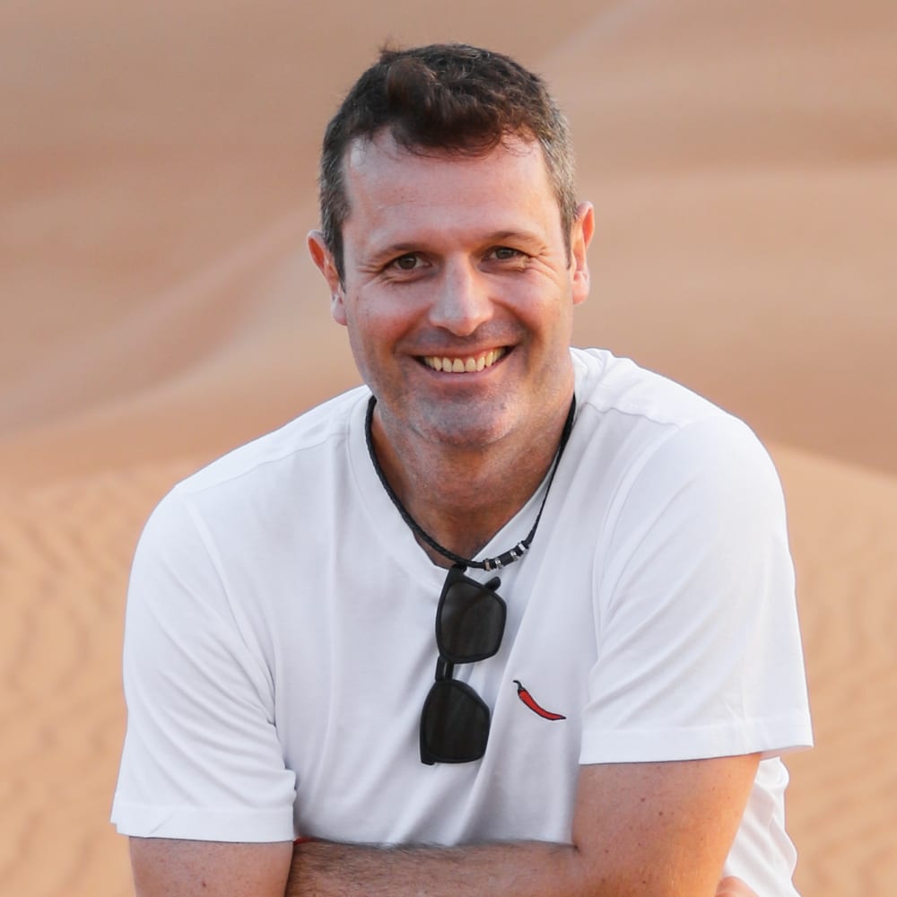 Ein lachender Mann mit kurzen dunklen Haaren, der ein weißes T-Shirt trägt und eine Sonnenbrille um den Hals hängen hat, steht in einer Wüstenlandschaft.