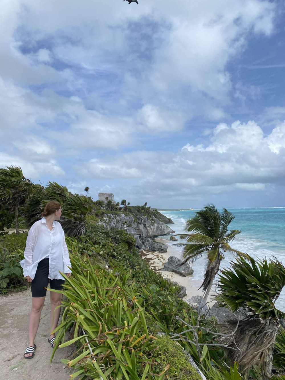 Eine Person mit weißem Hemd und schwarzer Shorts steht auf einem Küstenweg und blickt auf üppiges Grün, felsige Klippen, einen Sandstrand und türkisfarbenes Meer unter bewölktem Himmel in der Nähe von antiken Ruinen.