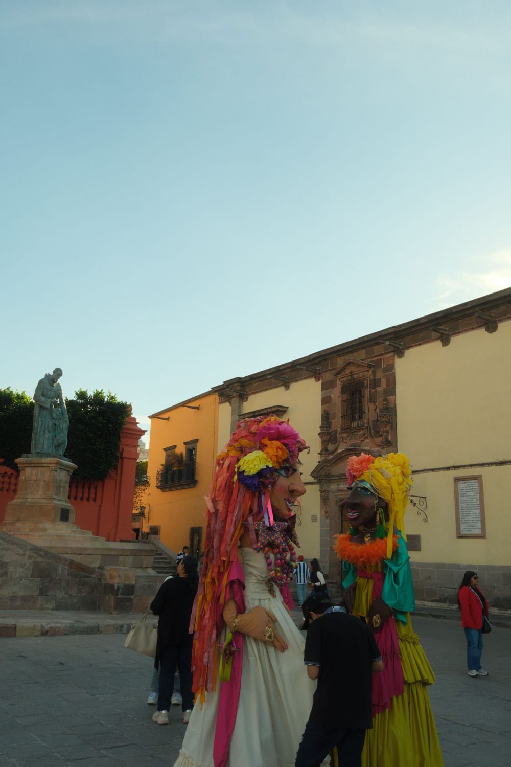 Zwei Personen in bunten traditionellen mexikanischen Riesen-Kopf-Kostümen auf einem historischen Platz mit kolonialen Gebäuden und einer Statue im Hintergrund bei Tageslicht.
