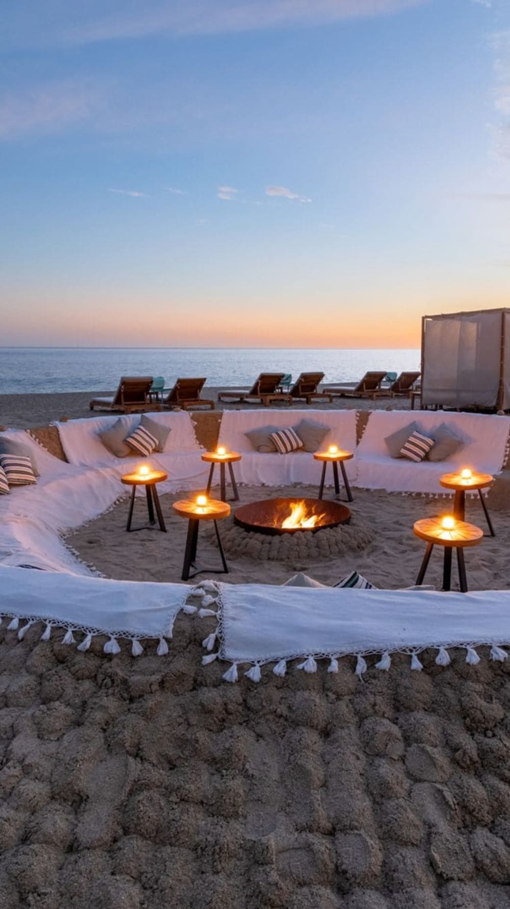 Gemütlicher runder Sitzbereich am Strand mit weißen Kissen, gestreiften Kissen, Holztischen mit Kerzen und einem zentralen Lagerfeuer vor ruhigem Ozean bei Sonnenuntergang.