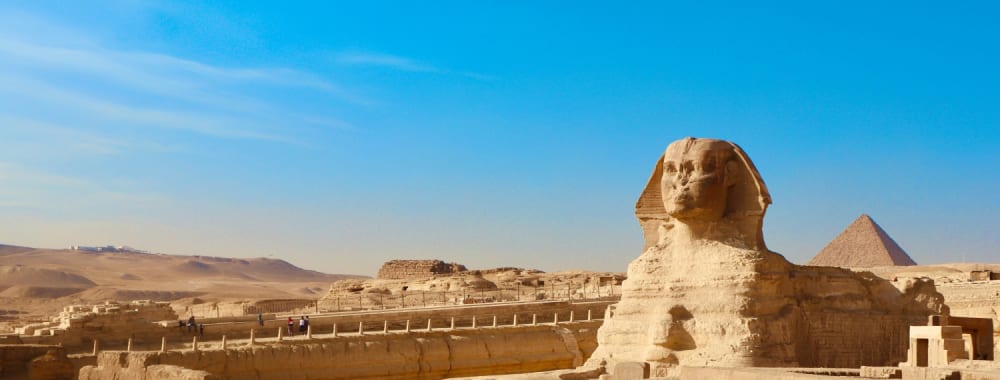 The Great Sphinx of Giza stands prominently against a clear blue sky, with the pyramids visible in the background under bright sunlight.