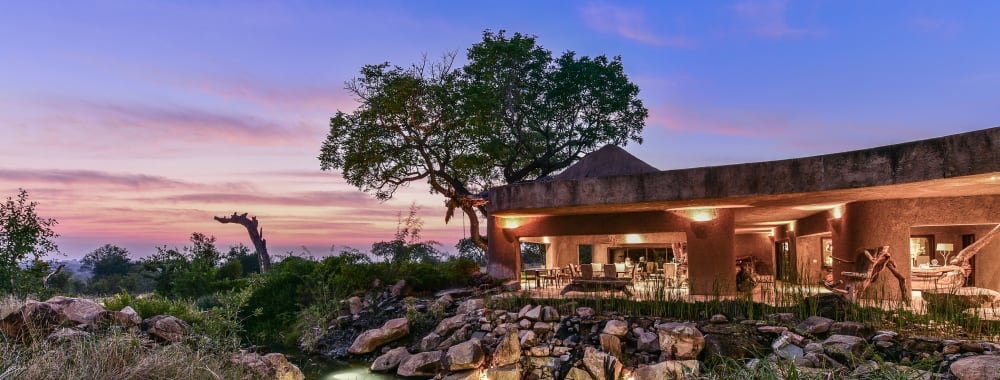 A luxurious lodge with an outdoor seating area, surrounded by rocks, water features, and lush greenery under a colorful sunset sky.