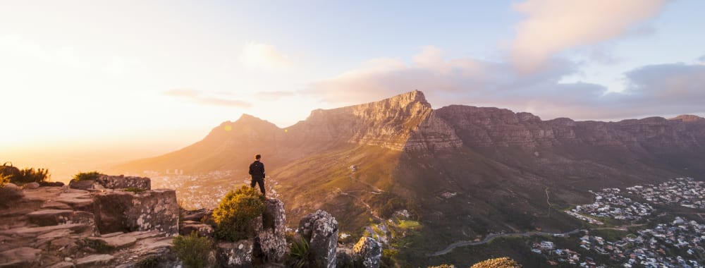 Eine Person steht auf einem felsigen Vorsprung und blickt auf eine Berglandschaft bei Sonnenuntergang.