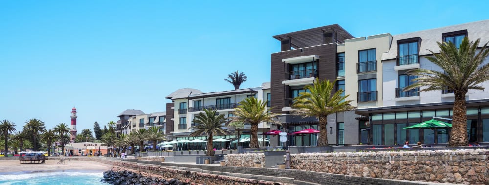 Modern hotel buildings along the waterfront with palm trees and outdoor seating areas under umbrellas.