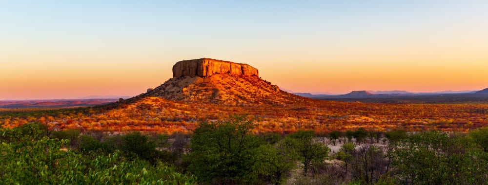 Ein flachgipfliger Berg, der vom Sonnenuntergang beleuchtet wird, mit klarem Himmel und spärlicher Vegetation im Vordergrund.