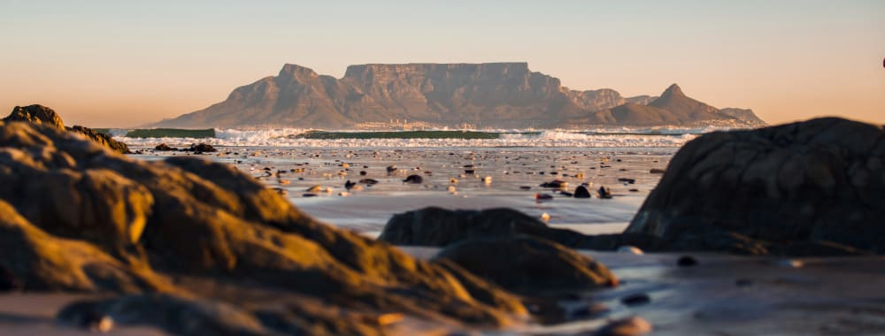 View of Table Mountain from a beach, with rocks and wet sand in the foreground during sunset.