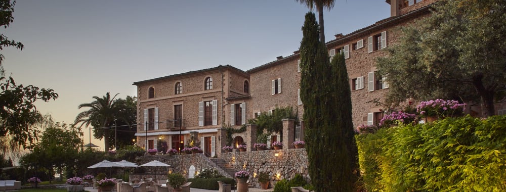 A large historic hotel with stone walls and multiple windows, surrounded by a well-maintained garden with trees, flowers, and outdoor seating areas at dusk.