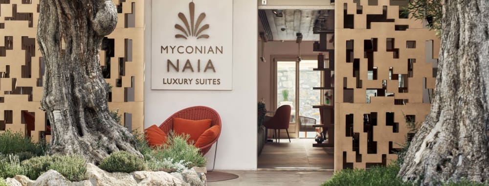Eingang zu Myconian Naia Luxury Suites umgeben von Bäumen und Felsen, mit einer modernen Dekorationswand und sichtbarem Innenraum durch die Tür.