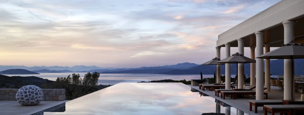 An infinity pool overlooking the sea at sunset with umbrellas and lounge chairs nearby.