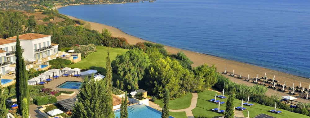 Vogelperspektive auf das Anassa Zypern Resort mit Schwimmbädern, üppigem Grün, Strand und Meer im Hintergrund.