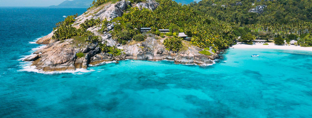 Eine tropische Insel mit üppigem Grün, felsigen Klippen und einem weißen Sandstrand, umgeben von türkisfarbenem Wasser.