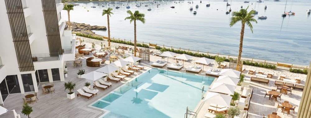 Luxurious hotel swimming pool with lounge chairs, umbrellas, and palm trees overlooking a marina in Ibiza, Spain.