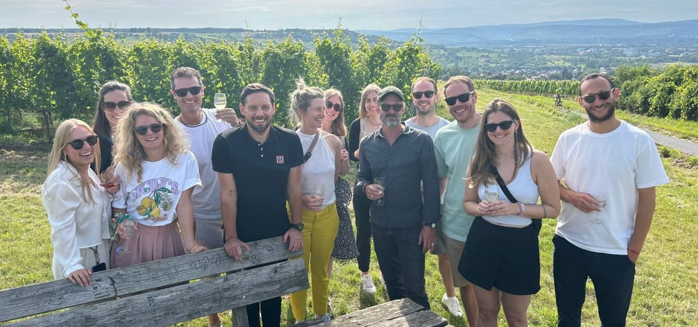 A group of fifteen people standing outdoors in a vineyard on a sunny day, some holding drinks and smiling.