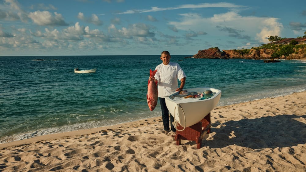 Koch steht an einem sandigen Strand neben einem Holzgestell mit frischem Fisch, hält einen großen roten Fisch, mit Meer und felsiger Küste im Hintergrund.