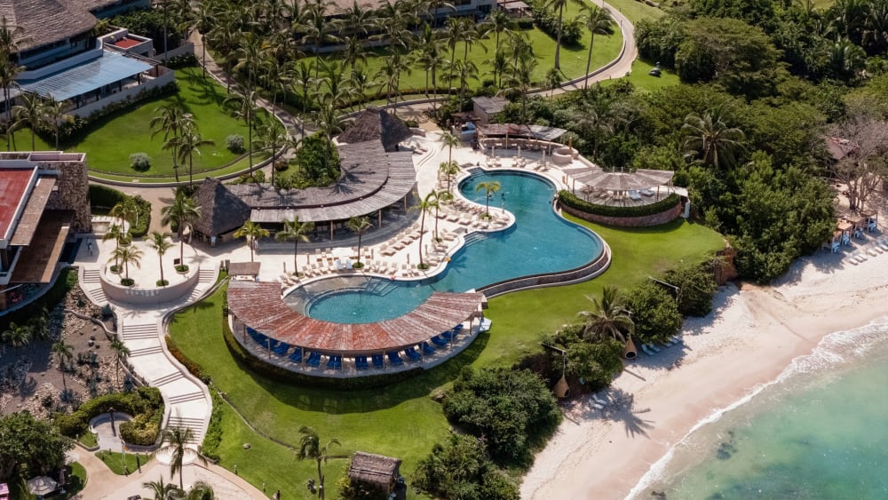 Luftaufnahme eines Resort-Anwesens mit einem einzigartig geformten Pool mit Liegestühlen, umgeben von Palmen, grünen Rasenflächen und angrenzend an einen Sandstrand mit klarem Meerwasser.