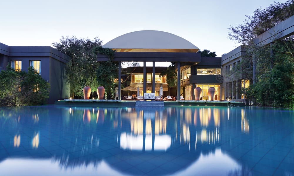 A luxurious resort or hotel with a large water body in the foreground, illuminated indoor spaces, and landscaped surroundings with trees and potted plants at dusk.