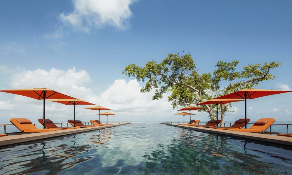 Ein ruhiger Außen-Infinity-Pool mit orangefarbenen Liegestühlen und Sonnenschirmen auf beiden Seiten, mit Blick auf einen weitläufigen bewölkten blauen Himmel und entfernten Horizont.