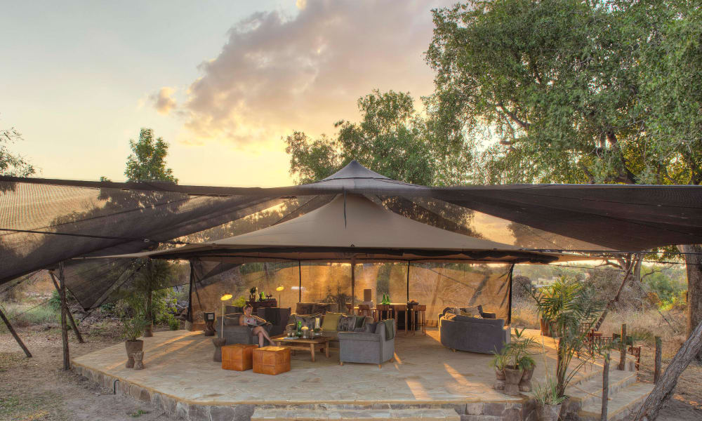 Ein luxuriöser Außen-Lounge-Bereich unter einer großen Überdachung mit Sofas, Stühlen und Tischen, umgeben von Bäumen bei Sonnenuntergang.
