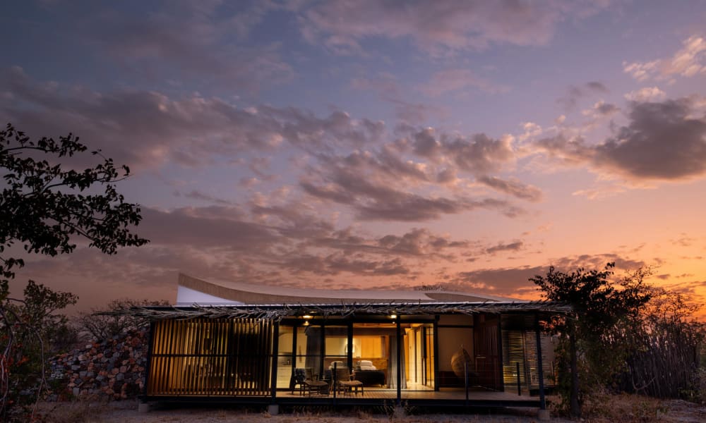 Eine moderne Suite mit Glasfront und warmem Innenlicht unter einem bunten, teils bewölkten Himmel bei Sonnenuntergang, umgeben von Natur.