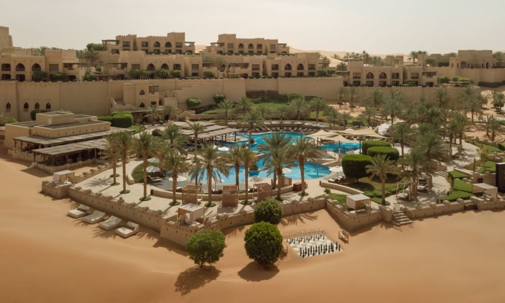 Ein luxuriöses Wüstenresort mit einem Pool, umgeben von Palmen und Sonnenliegen, in einer trockenen Wüstenumgebung.