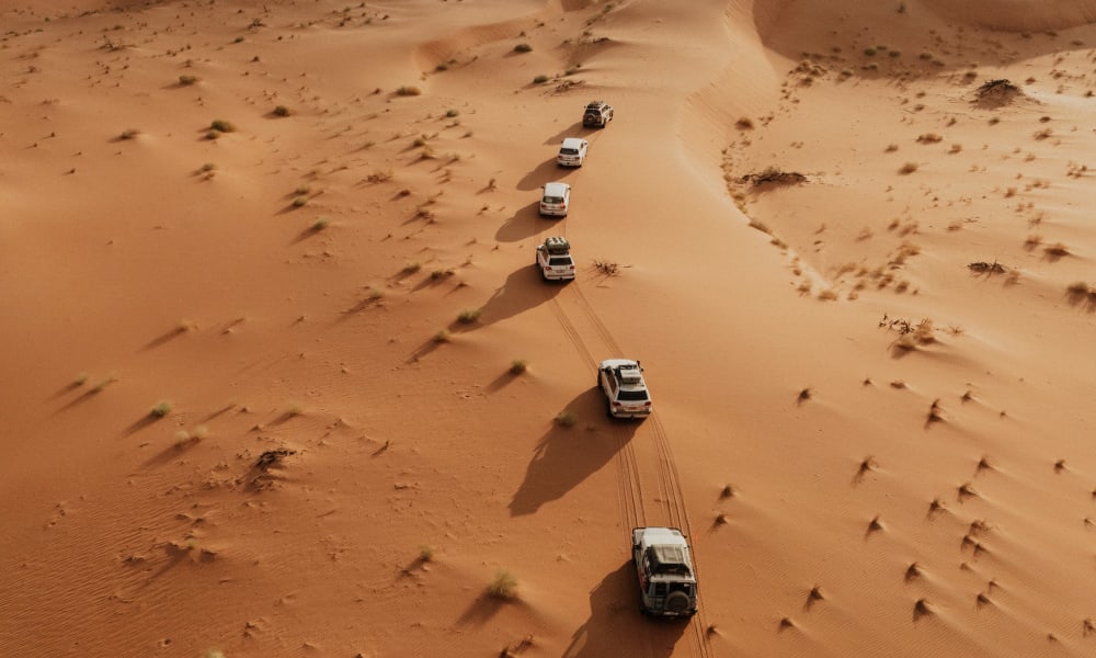 Seven SUVs drive in a row through the sand dunes of the desert, with long shadows cast by the sun on the left side of the vehicles.