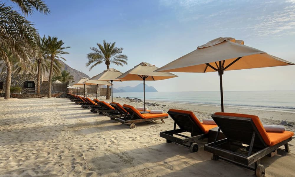 Eine Reihe leerer Liegestühle mit orangefarbenen Kissen und großen Sonnenschirmen am sandigen Strand, mit Palmen und Bergen im Hintergrund.