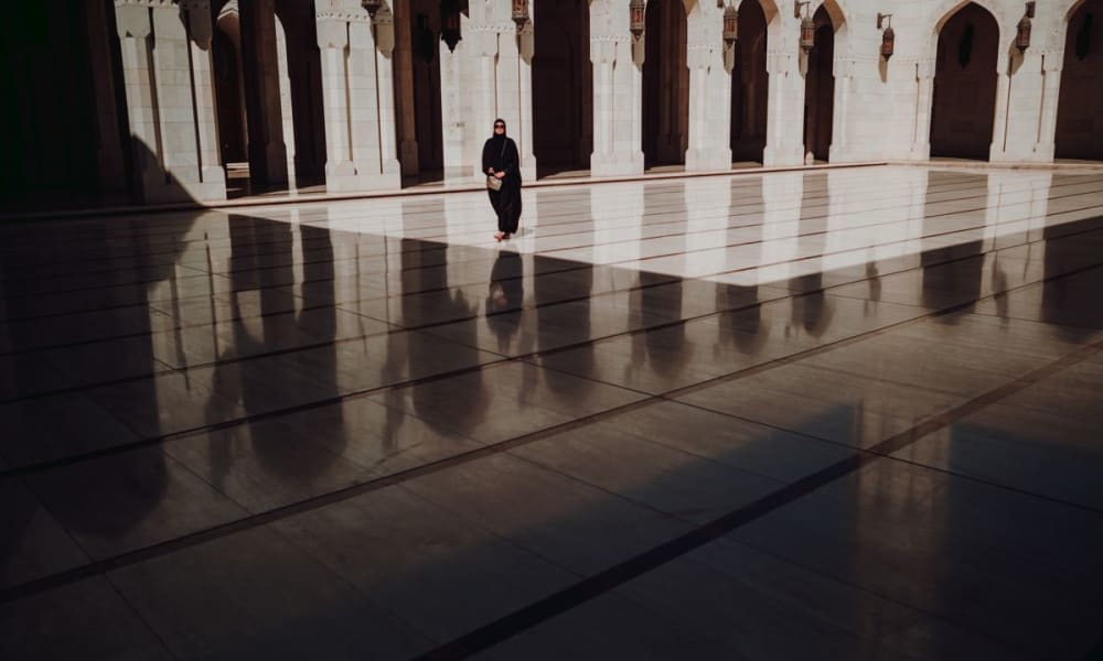 A solitary person dressed in black walking in an arched, sunlit courtyard with intricate shadows on the polished floor.